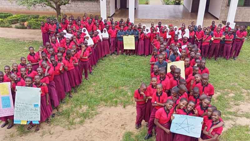 Un grand groupe de jeunes élèves en uniforme rouge devant le bâtiment de l'école, formant une grande lettre Pi.